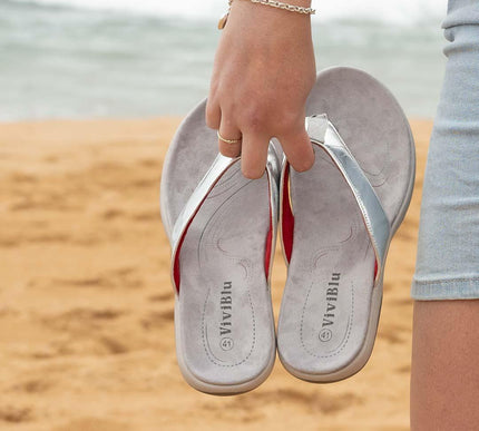 Person holding silver and red flip flops on sandy beach with ocean in background
