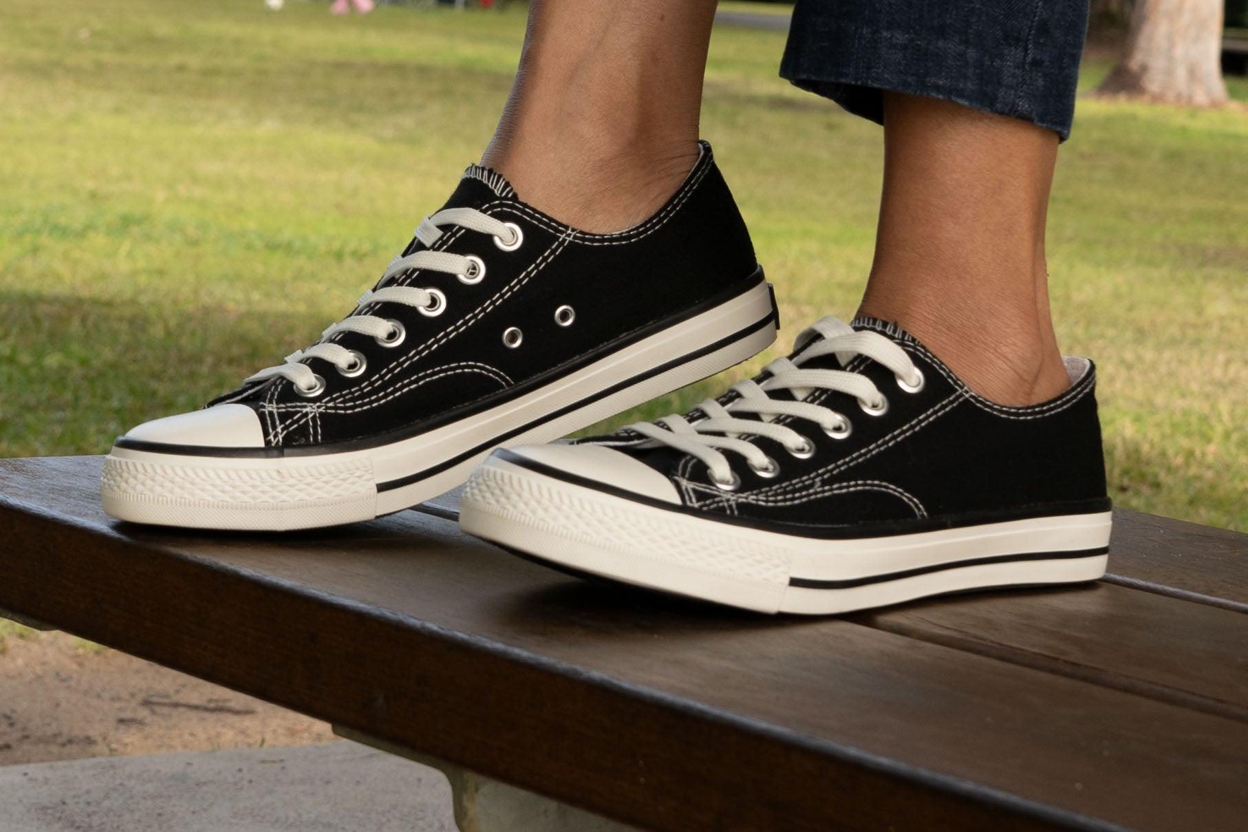 Black canvas low-top sneakers with white soles and laces worn outdoors on a bench